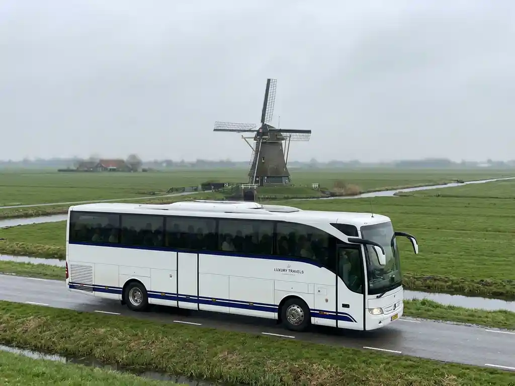 Witte luxe touringcar rijdt over een Nederlandse polderwegen met groene weilanden en een windmolen op de achtergrond.