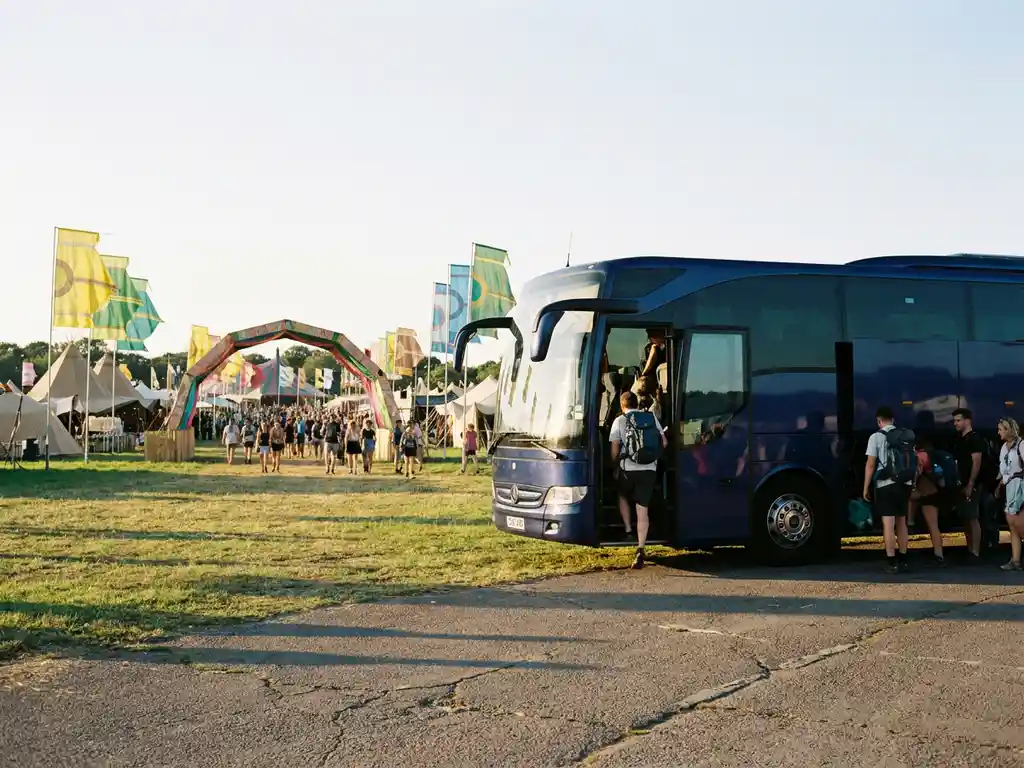 Donkerblauwe touringcar bij festivalingang met uitstappende passagiers, kleurrijke tenten en vlaggen op de achtergrond in middagzon.