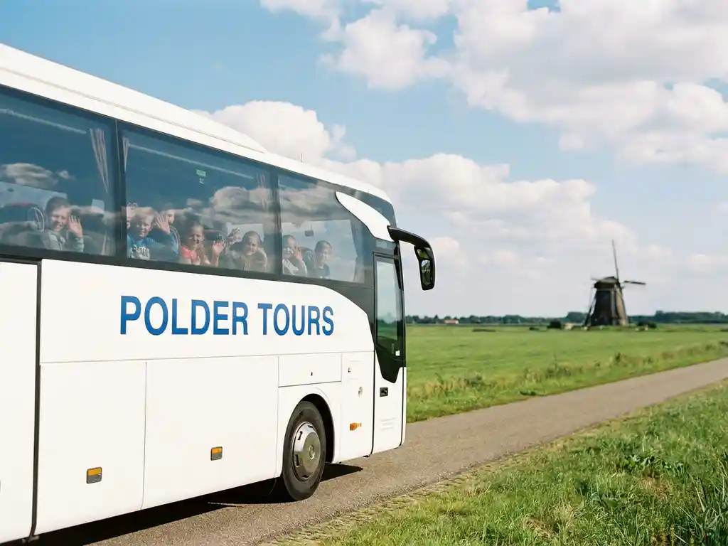 Witte touringcar met schoolkinderen op een polderweg, met groene weilanden en een windmolen op de achtergrond.