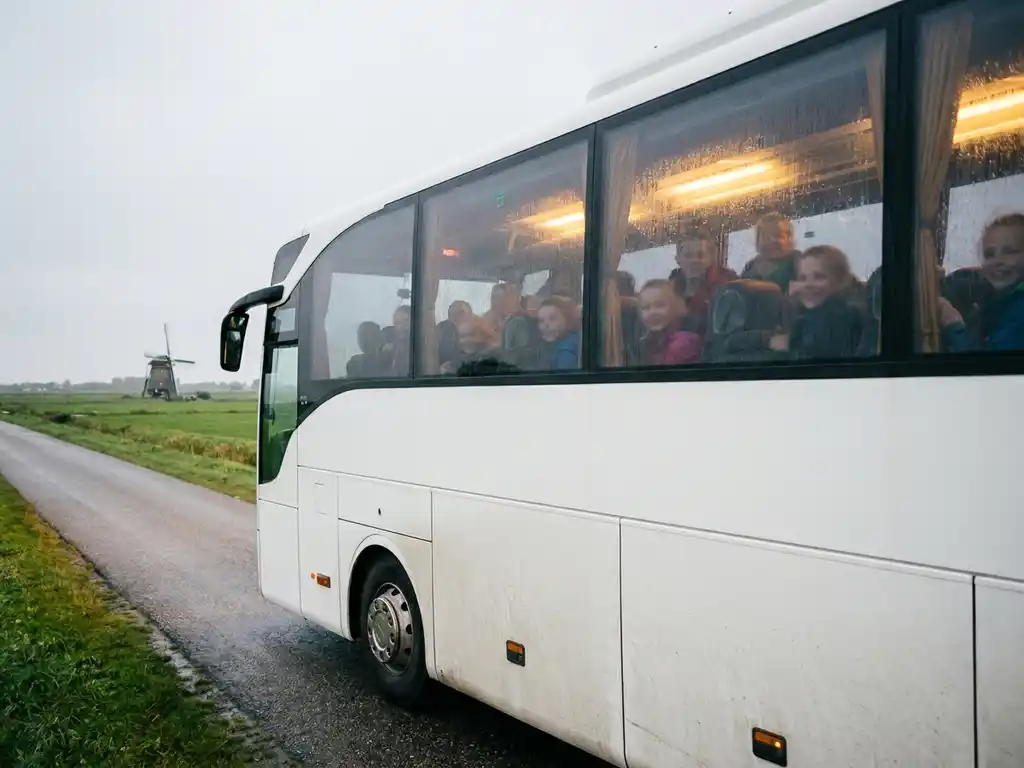 Moderne touringcar met schoolkinderen rijdt over een Nederlandse polderwegweg, met groene weilanden en een windmolen op de achtergrond.