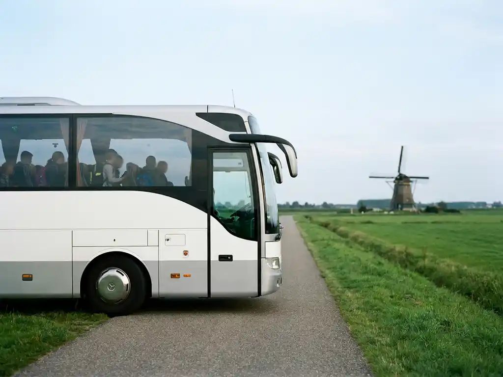 Witte touringcar met schoolkinderen rijdt over een Nederlandse polderwegen, met groene weilanden en een windmolen op de achtergrond.
