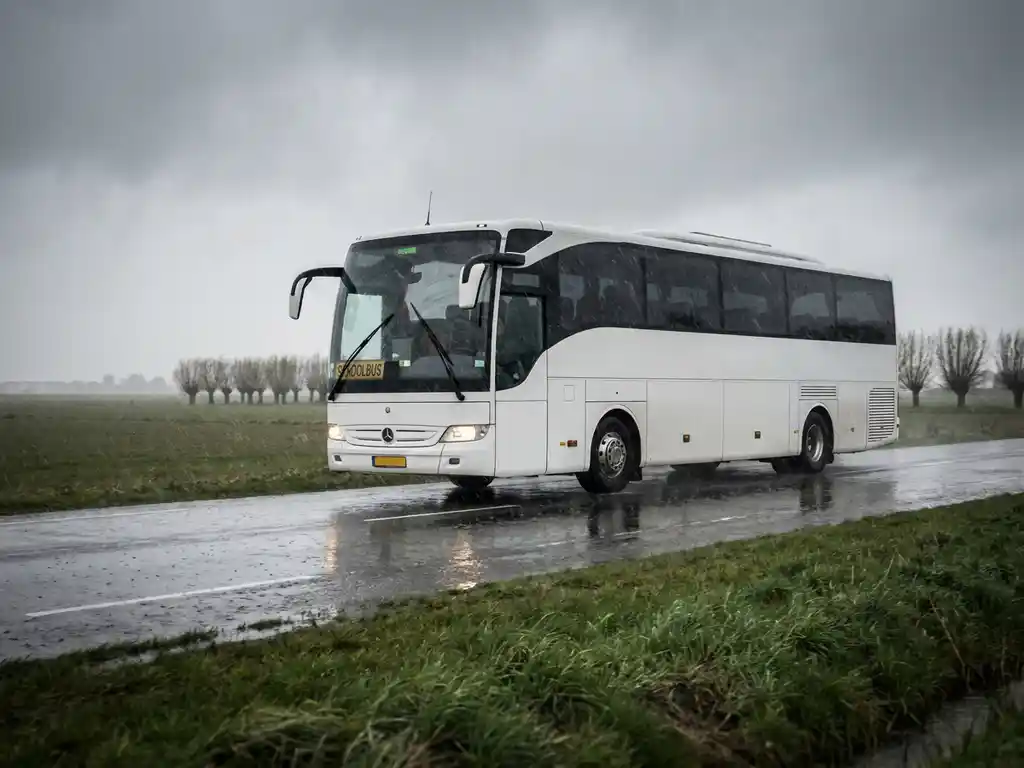 Touringcar met schoolkinderen rijdt door regenachtig Nederlands polderlandschap met grijze bewolkte lucht en natte asfaltweg.