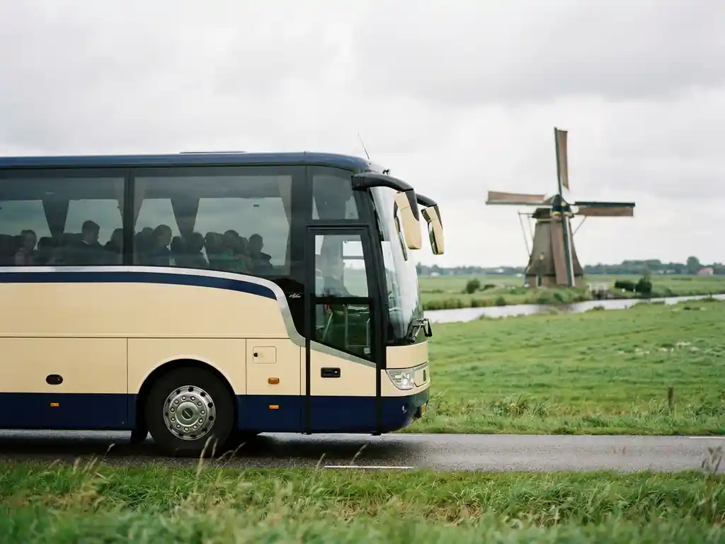 Touringcar met schoolkinderen rijdt over een polderweg met groene weilanden en een windmolen op de achtergrond.