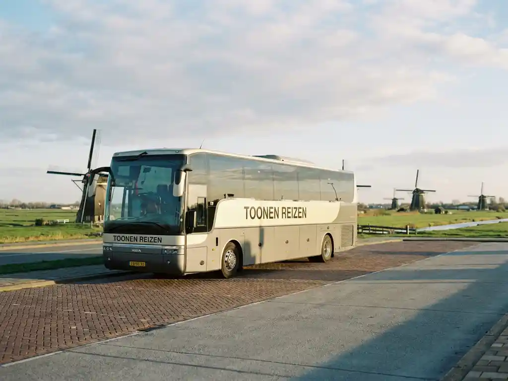 Touringcar van Toonen Reizen geparkeerd op een Nederlandse bushalte, met windmolens en een vlak polderlandschap op de achtergrond.