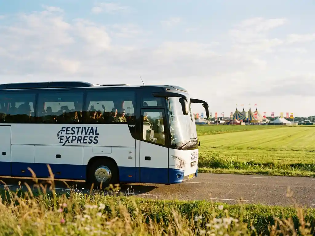 Touringcar met festivalgangers rijdt door een zonnig Nederlands polderlandschap, met kleurrijke festivaltenten op de achtergrond.