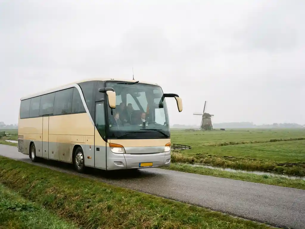 Moderne touringcar rijdt door vlak Nederlands polderlandschap met windmolen op de achtergrond onder bewolkte lucht.
