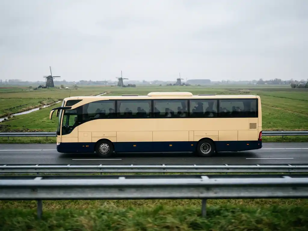 Touringcar rijdt over Nederlandse snelweg met vlak polderlandschap, windmolens en bewolkte lucht op de achtergrond.