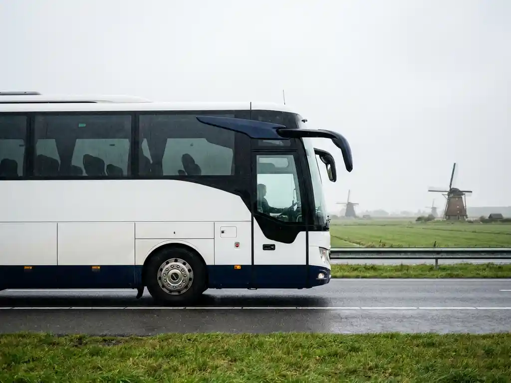 Witte en marineblauwe touringcar rijdt over Nederlandse snelweg langs groene polders en windmolens onder bewolkte lucht.
