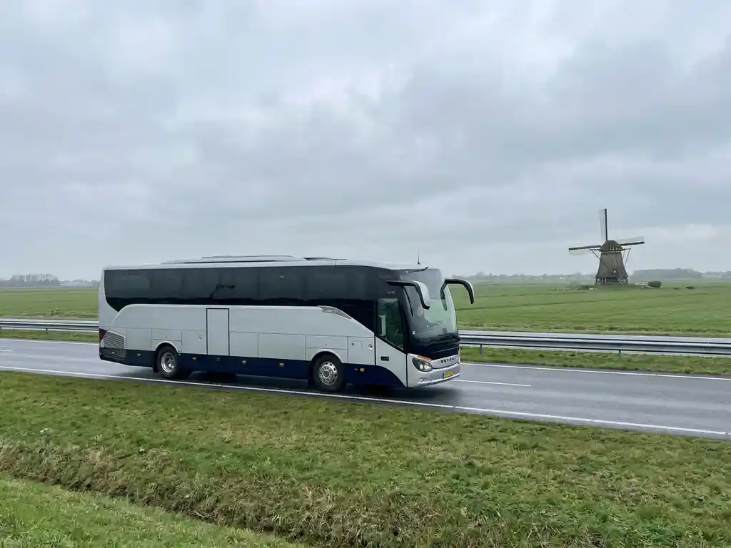 Moderne touringcar op Nederlandse snelweg met groene polders en windmolen op de achtergrond onder bewolkte lucht.