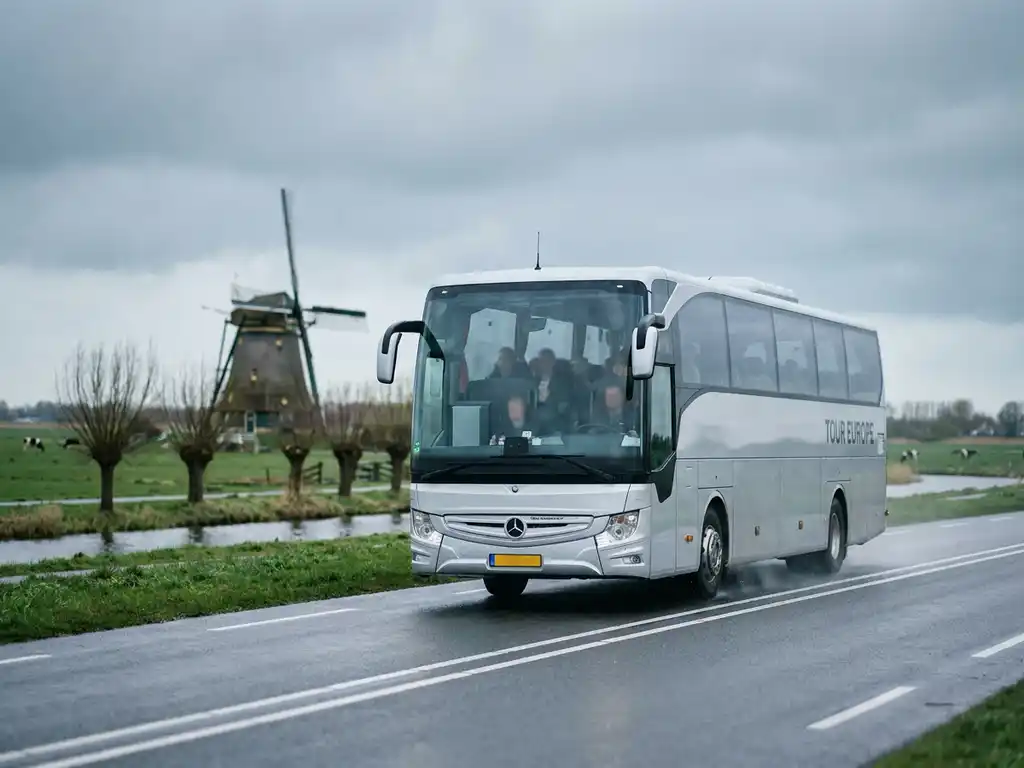 Zilverwitte touringcar op Nederlandse snelweg met groene polders en windmolen op de achtergrond.
