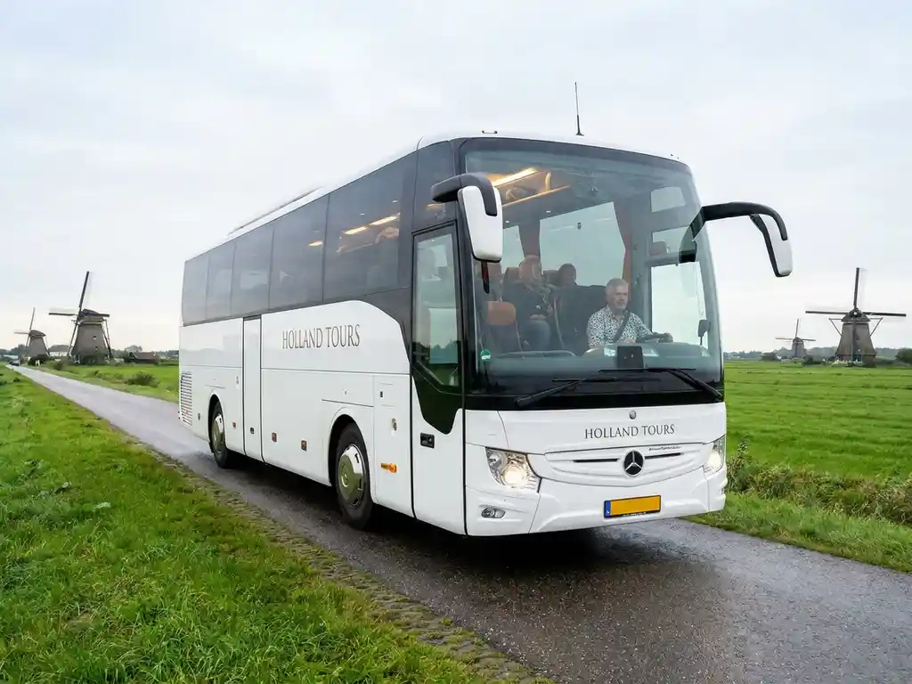 Moderne touringcar met passagiers rijdt door Nederlands polderlandschap met windmolens en groene weilanden onder bewolkte lucht.