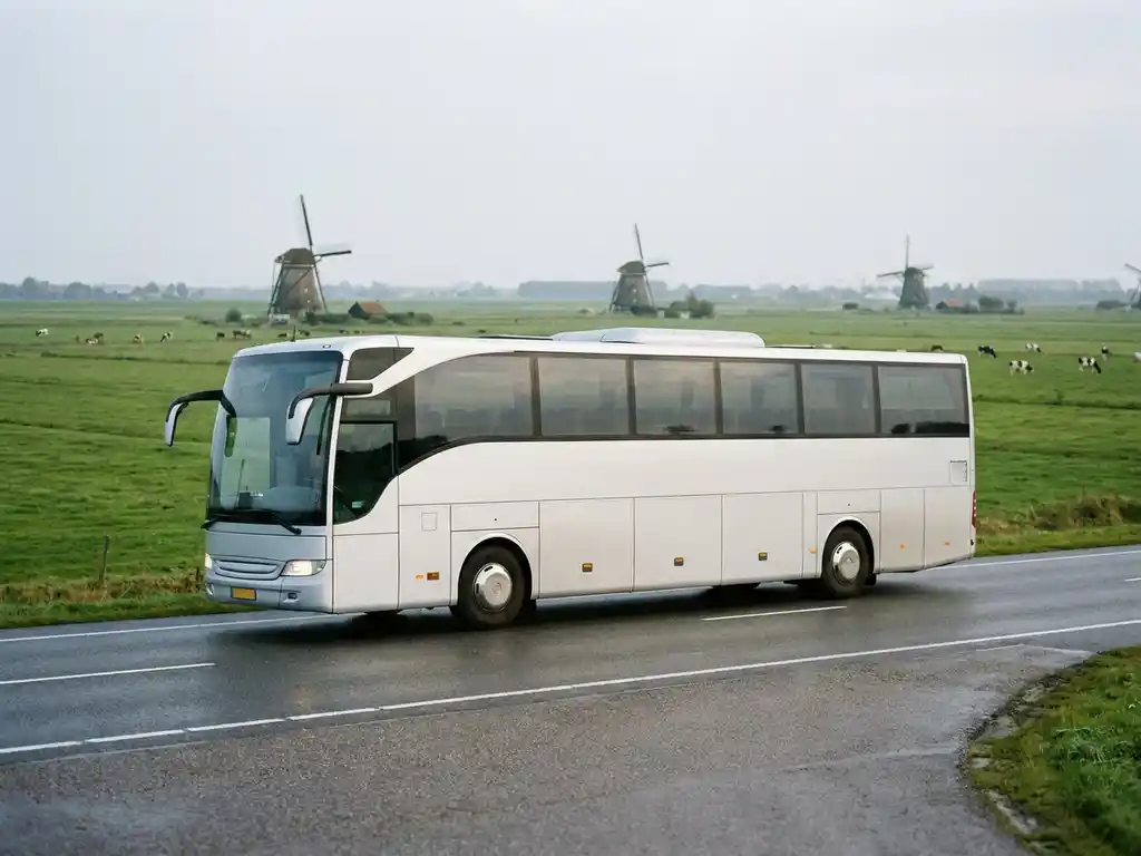 Moderne touringcar op Nederlandse snelweg met windmolens en groene polders op de achtergrond onder bewolkte lucht.