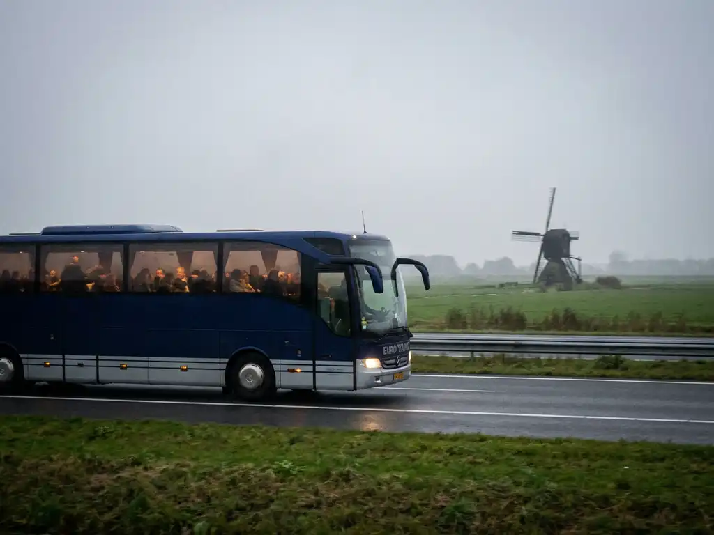 Moderne touringcar rijdt over Nederlandse snelweg door vlak polderlandschap met windmolen op de achtergrond.