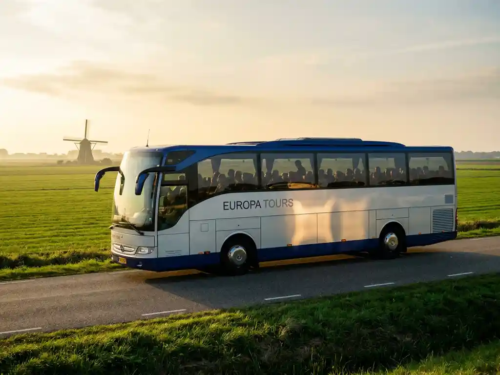 Moderne touringcar rijdt door Nederlands polderland met groene weiden, windmolen op achtergrond en gouden middaglicht.