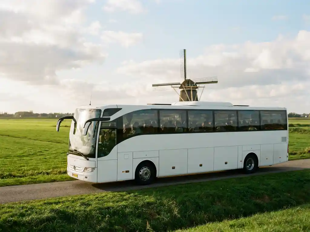 Touringcar rijdt over een Nederlandse polderdijk met groene weilanden, een windmolen op de achtergrond en bewolkte lucht.