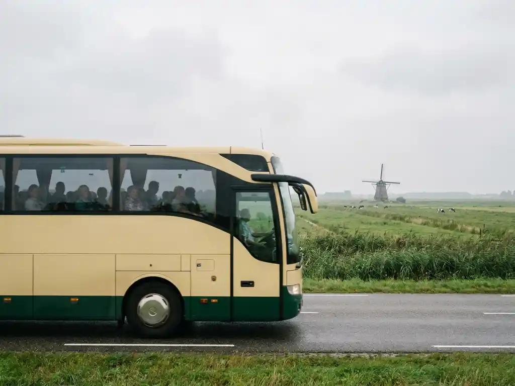 Moderne touringcar met passagiers rijdt door Nederlands polderlandschap met windmolen en groene weilanden op de achtergrond.