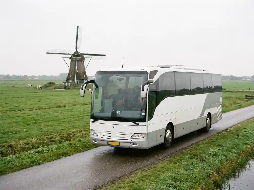 Moderne touringcar voor 50 passagiers rijdt over een Nederlandse polderwweg met groene weilanden en een windmolen op de achtergrond.