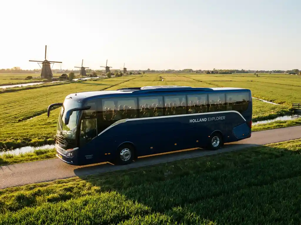 Touringcar rijdt door een Nederlands polderlandschap met windmolens en groene weiden in warm middaglicht.