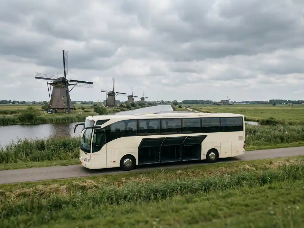 Touringcar met open bagageruimten rijdt door Nederlands polderlandschap met windmolens en groene weilanden.