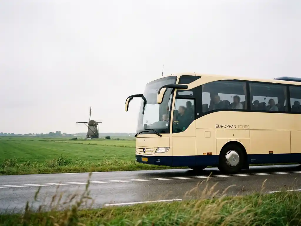 Moderne touringcar in crème en marineblauw rijdt over een polderweg met groene weilanden en een windmolen op de achtergrond.