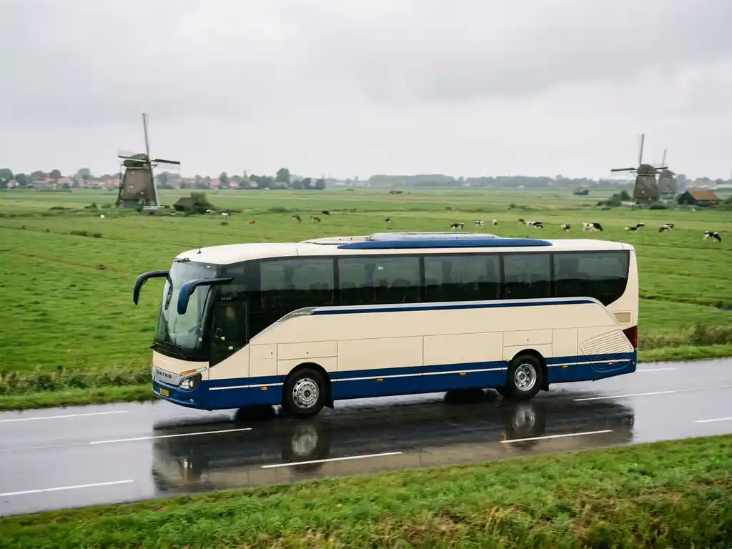 Moderne touringcar rijdt door Nederlands polderlandschap met windmolens en groene weilanden onder bewolkte lucht.