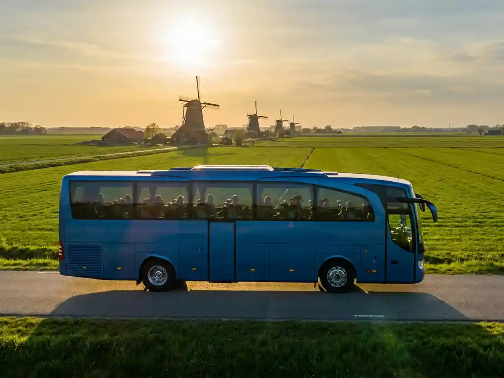 Moderne touringcar met studenten rijdt door een vlak Nederlands polderlandschap met windmolens en groene weiden in gouden middaglicht.