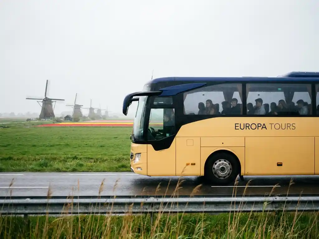 Moderne touringcar met studenten rijdt door een Hollands polderlandschap met windmolens en tulpenvelden op de achtergrond.