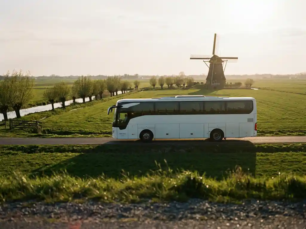Witte touringcar met sportgroep rijdt door Nederlands polderlandschap met groene weilanden en windmolen op de achtergrond.