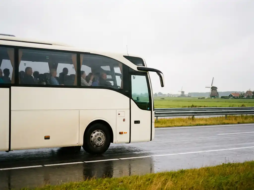 Witte touringcar met passagiers op Nederlandse snelweg, omgeven door groene polders en windmolens onder bewolkte lucht.