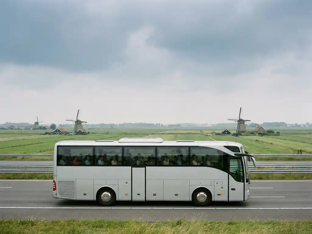 Touringcar met passagiers op Nederlandse snelweg, omgeven door polderlandschap met windmolens onder bewolkte lucht.
