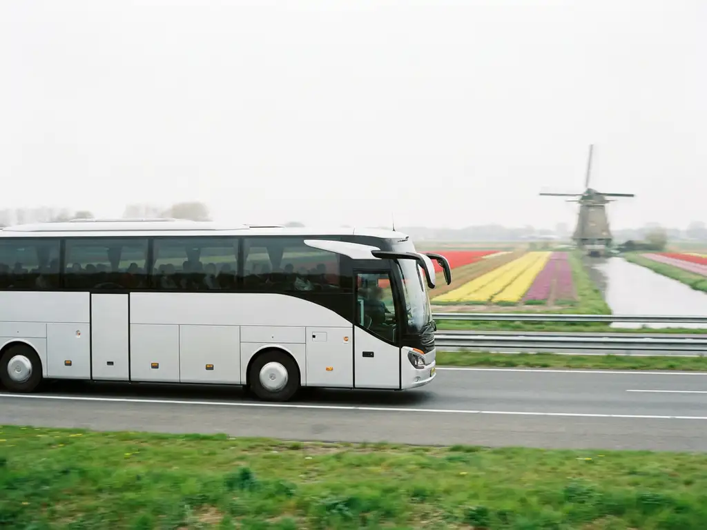 Moderne touringcar rijdt over Nederlandse snelweg langs tulpenvelden en een windmolen onder bewolkte lucht.