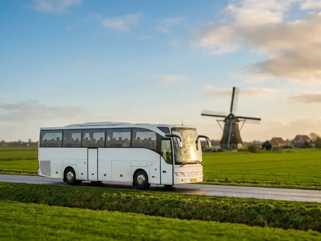 Witte touringcar met vijftig passagiers op een Nederlandse polderwegen, groene weilanden en molen op de achtergrond in warm middaglicht.