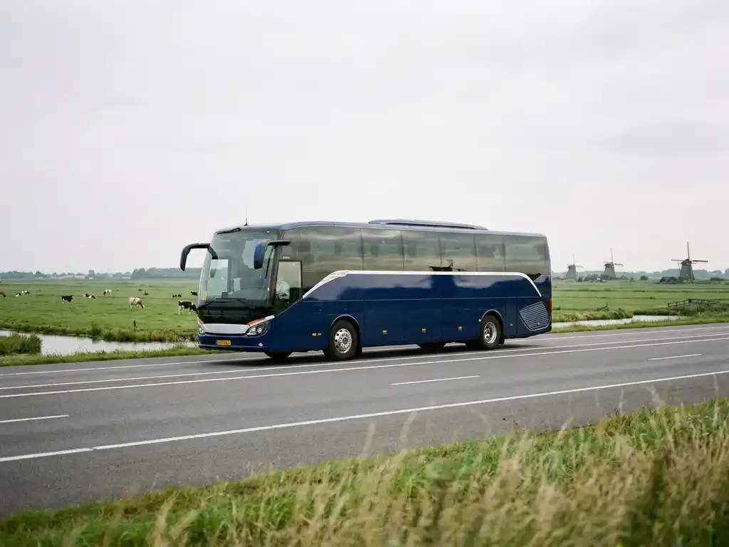 Moderne touringcar op Nederlandse snelweg met groene polders en windmolens op de achtergrond in bewolkt daglicht.